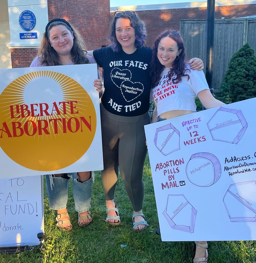 Three people standing with their arms around each other holding signs that say "liberate abortion" and "abortion pills by mail."